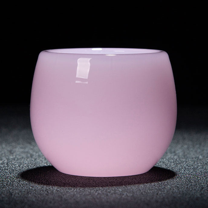 A small translucent pink glass cup with a rounded shape, shown on a dark textured surface against a black background. A white label is visible on the upper left side of the cup.