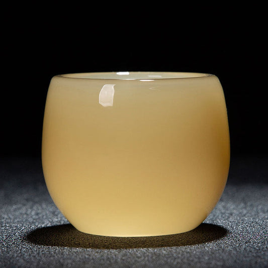 A small round translucent ceramic cup with a pale yellow-beige tint, shown on a dark textured surface against a black background. A white label is visible on the upper left side of the cup.