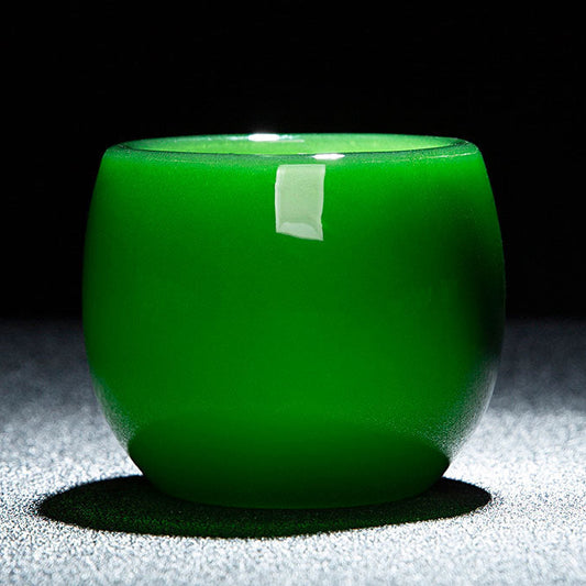 A small round green glass cup with a glossy finish, shown on a dark background with a gray textured surface beneath it. A white label is attached to the front.