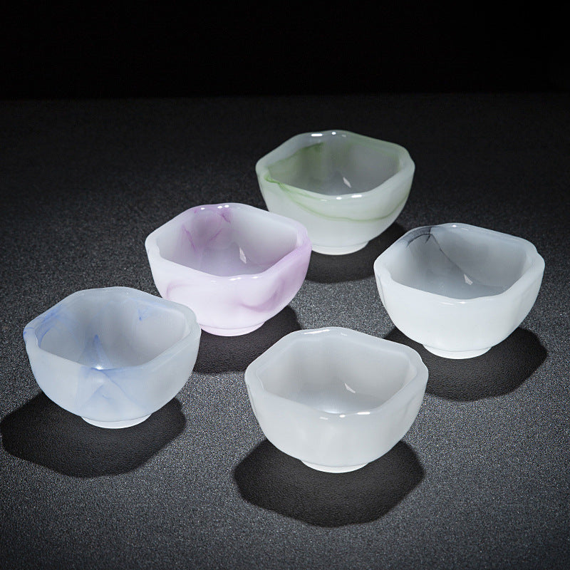 Five small translucent glass cups in assorted pastel colors, including white, light purple, pale blue, and pale green, arranged on a dark textured surface.
