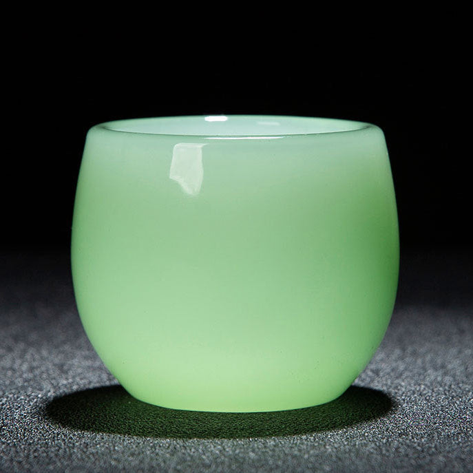 A small translucent jade-green glass cup with a rounded shape, shown on a dark textured surface against a black background. A red banner at the bottom contains Chinese text with product dimensions and capacity.